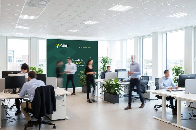 Viele Schreibtischen und Mitarbeitern in hellem Büroraum - SAKD Banner im Hintergrund