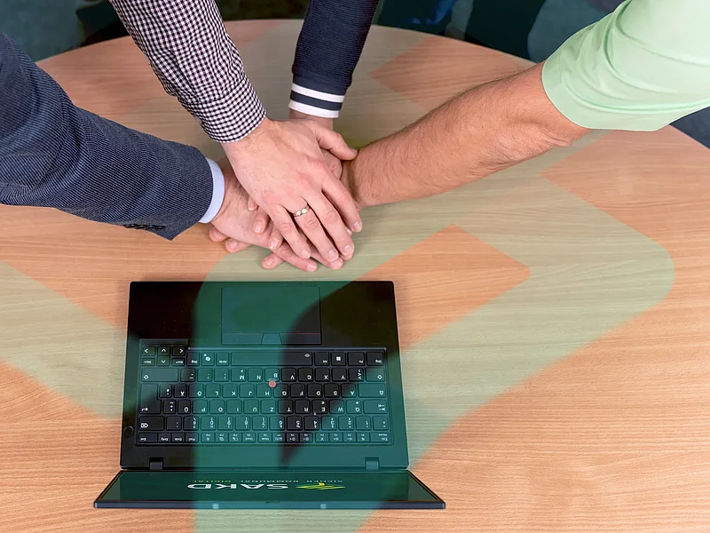 Mehrere Hände liegen als Zeichen des Teamgeists übereinander auf einem Tisch neben einem Laptop.