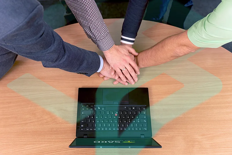 Mehrere Hände liegen als Zeichen des Teamgeists übereinander auf einem Tisch neben einem Laptop.