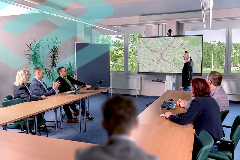 Ein Mitarbeiter zeigt im Seminarraum auf eine digitale Stadtkarte auf einem großen Bildschirm, das Team hört zu.