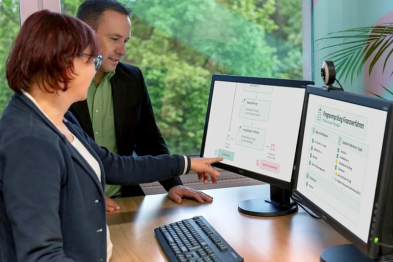 Eine Mitarbeiterin und ein Mitarbeiter prüfen an zwei Monitoren den Ablauf einer Programmprüfung für Finanzverfahren.