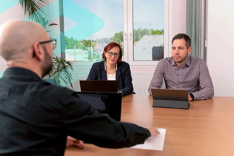 Ein Mitarbeiter legt einer Kollegin und einem Kollegen am Besprechungstisch ein Dokument vor.